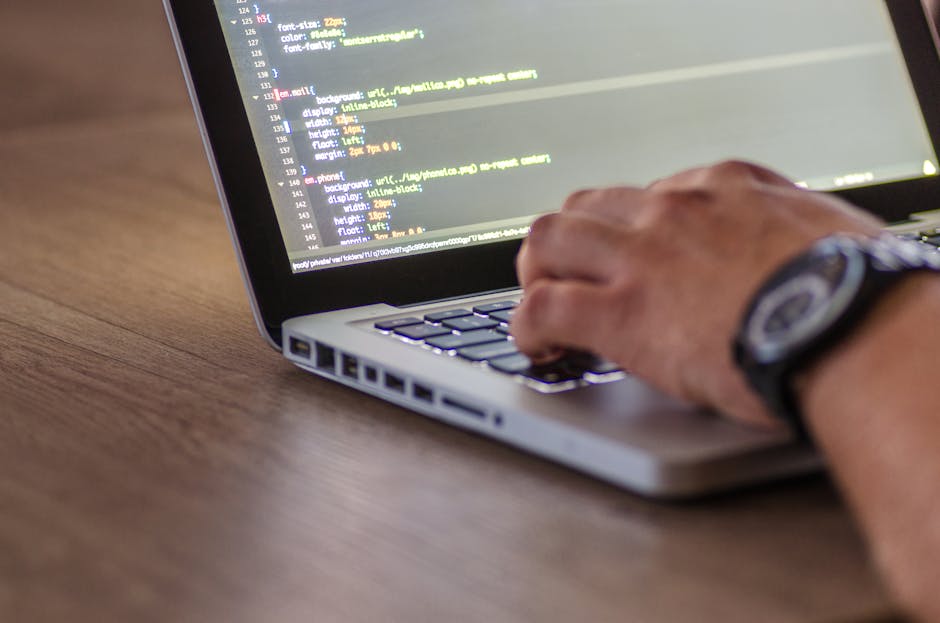 A close-up of a person's hand typing on a laptop keyboard, with the screen displaying lines of code in a text editor. The individual is wearing a dark wristwatch. The laptop is placed on a wooden surf