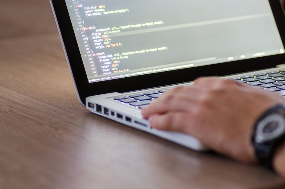A close-up of a person's hand typing on a laptop keyboard, with the screen displaying lines of code in a text editor. The individual is wearing a dark wristwatch. The laptop is placed on a wooden surf